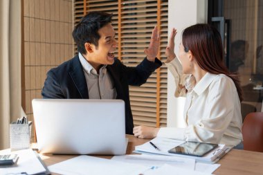 Resmi giyinmiş erkek ve kadın işadamları ofiste dizüstü bilgisayar ve belgelerle birlikte oturuyor ve birlikte çalışıyorlar. Birbirlerini kutluyorlar ve tezahürat yapıyorlar. Heyecanlarının ifadesi olarak. Bitirdikten ve başardıktan sonra.