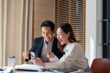 Resmi giyinmiş erkek ve kadın işadamları ofiste belgelerle birlikte bir masada oturuyor ve birlikte çalışıyorlar. İyi haber ya da başarı bilgisi aldıktan sonraki heyecanları için birbirlerini kutluyor ve alkışlıyorlar. 