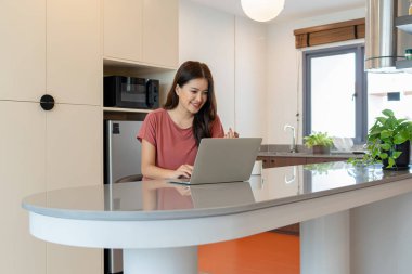 Pembe tişörtlü mutlu bir Asyalı kadın, modern bir mutfaktaki bir bilgisayarda uzaktan iletişimin tadını çıkarırken gülümsüyor ve jestler yapıyor..