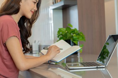 Gülümseyen Asyalı bir kadın mutfak tezgahında dizüstü bilgisayarının yanına mavi bir defter yazar. Uzak bir çalışma sırasında düzenli ve üretken kalır..