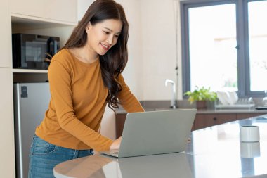 Gündelik kahverengi gömlekli neşeli bir Asyalı kadın, rahat evindeki modern bir mutfağın tezgahında duruyor, gülümseyen bir dizüstü bilgisayarla çalışıyor ya da internette geziniyor..
