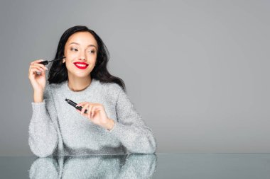 Kırmızı dudaklı, mutlu esmer kadın maskarayı tutarken, bakışlarını griye çeviriyor.