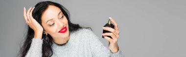pleased woman with red lips holding bottle with perfume isolated on grey, banner clipart