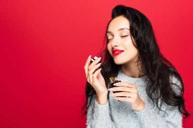 young pleased woman in sweater holding bottle and smelling perfume isolated on red clipart