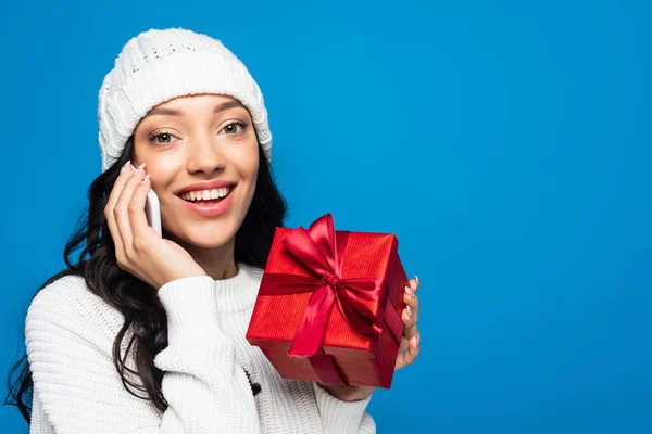 ニット帽をかぶった幸せな女性がプレゼントを包み青で隔離されたスマートフォンで話しています — ストック写真
