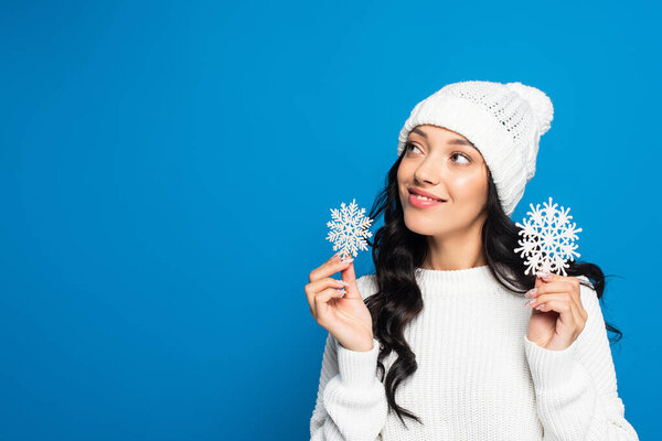 joyful woman in knitted hat holding decorative snowflakes isolated on blue