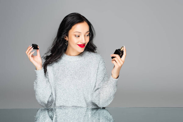 happy woman with red lips looking at bottle with perfume isolated on grey 