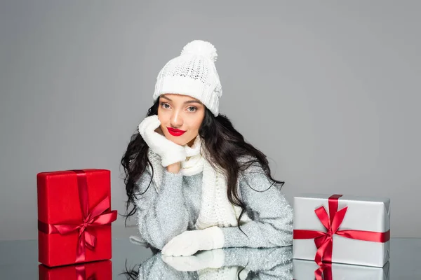 brunette woman in winter outfit looking at camera near presents isolated on grey