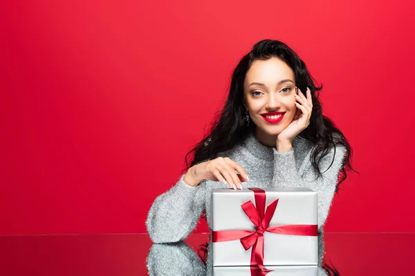 赤い上に孤立したプレゼントを包んだカメラを見てセーターを着た若い幸せな女性 — ストック写真