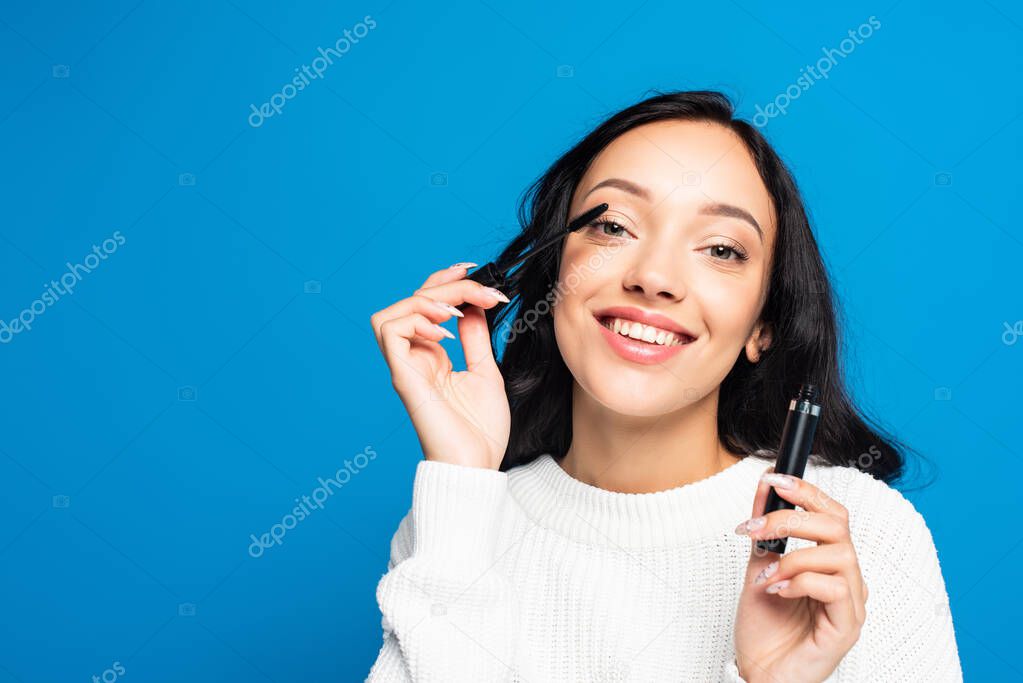 happy brunette woman applying mascara isolated on blue