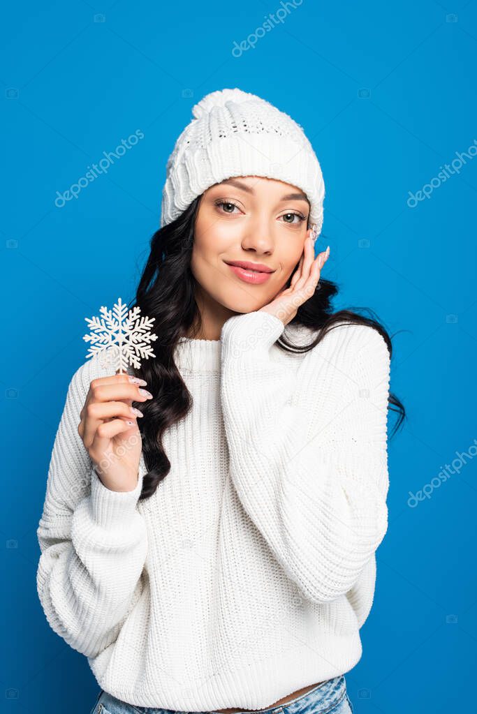 pleased woman in knitted hat holding decorative snowflake isolated on blue