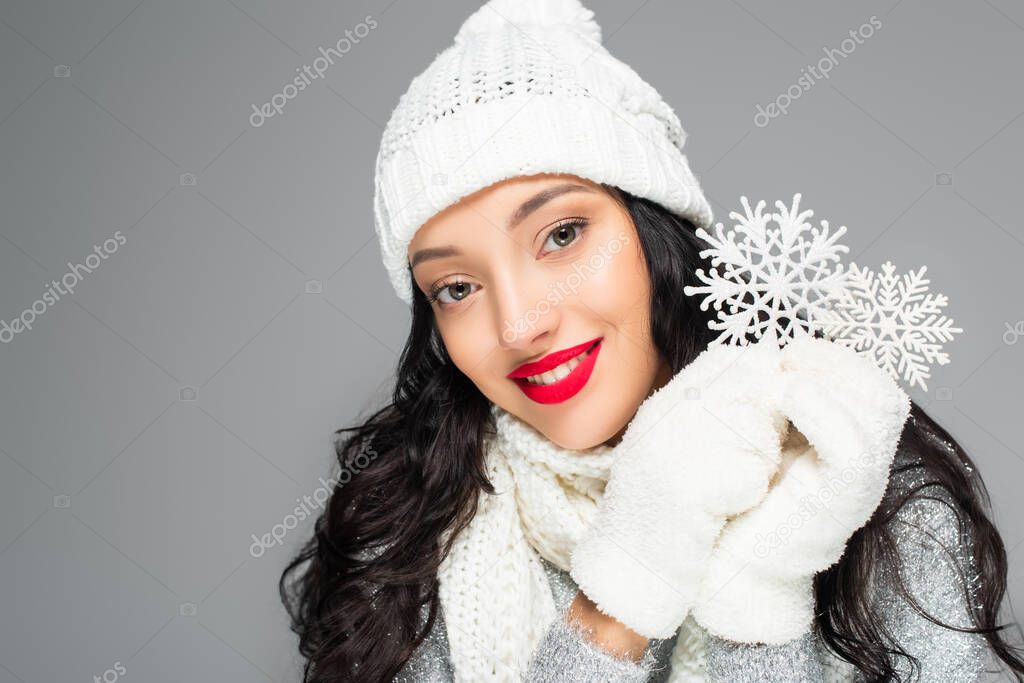 joyful woman in winter outfit looking at camera and holding decorative snowflakes isolated on grey
