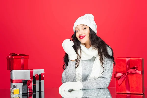 joyful woman in winter outfit holding lipstick near christmas presents and bottles with perfume isolated on red
