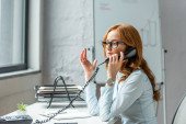 Rothaarige Geschäftsfrau gestikuliert und telefoniert am Festnetztelefon, während sie am Arbeitsplatz vor verschwommenem Hintergrund sitzt