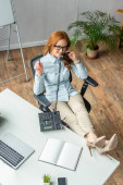 Hochwinkelaufnahme einer lächelnden Geschäftsfrau mit überkreuzten Beinen, die am Festnetztelefon in der Nähe ihres Arbeitsplatzes mit Geräten spricht