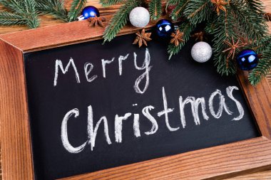 Kahverengi ahşap arka planda çam dalları, takılar ve anason yıldızlarının yanındaki mutlu noel harfleriyle karatahtaya yakından bakın.