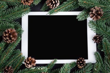 Dijital tablet, ladin dalları ve siyah zemin üzerinde çam kozalakları ile düz yatıyordu, yeni yıl konsepti