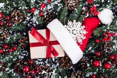 Yapay karlı ladin dalları, Noel Baba şapkası yanında hediye kutusu ve siyah arka planda dekoratif kar tanesi.