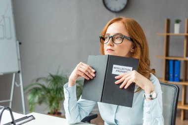 Thoughtful lawyer holding book with intellectual property lettering, while looking away at workplace on blurred background clipart