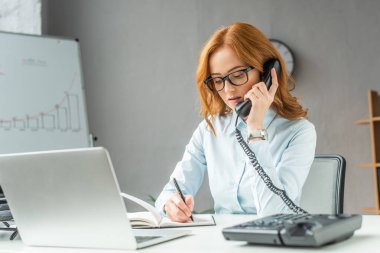 Sabit hattan konuşan kızıl saçlı iş kadını, iş yerindeki defterine bulanık ön planda yazı yazarken,