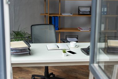 Ofis içi masa, ofis koltuğu ve ön planda bulanık pencereli raflar