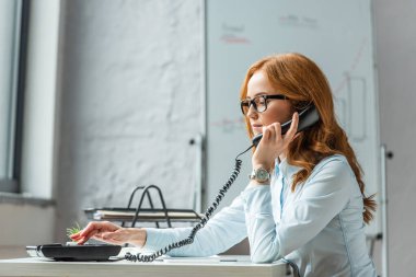 Focused businesswoman with handset dialing number on landline telephone, while sitting at workplace on blurred background clipart