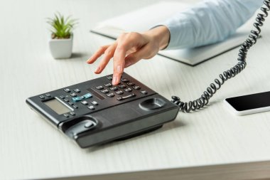 Cropped view of businesswoman dialing number on landline telephone on table with notebook and plant  clipart