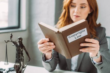 Concentrated redhead lawyer reading book with copyright law lettering near themis figurine with blurred window on background clipart