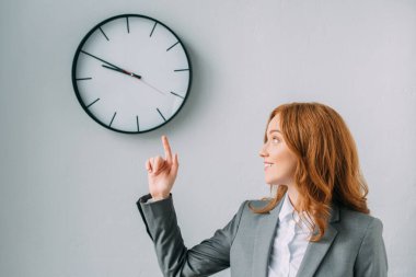 Smiling redhead businesswoman pointing with finger and looking at wall clock on grey clipart