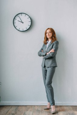 Full length of confident businesswoman with crossed arms standing near wall clock in office clipart