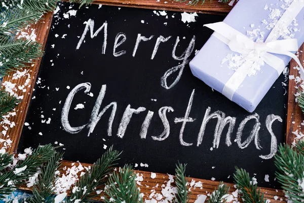 stock image Close up view of chalkboard with merry christmas lettering, little present, pine branches and artificial snow