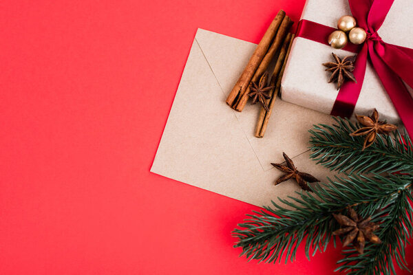Top view of gift with envelope, cinnamon sticks, anise stars and pine branch on red background