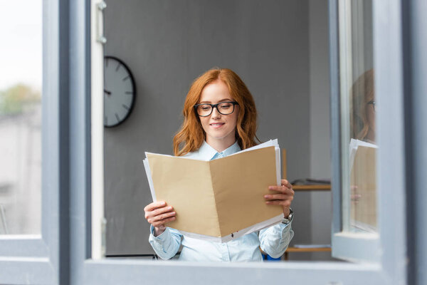 Smiling businesswoman looking at folder with paper sheets in office with blurred window on foreground