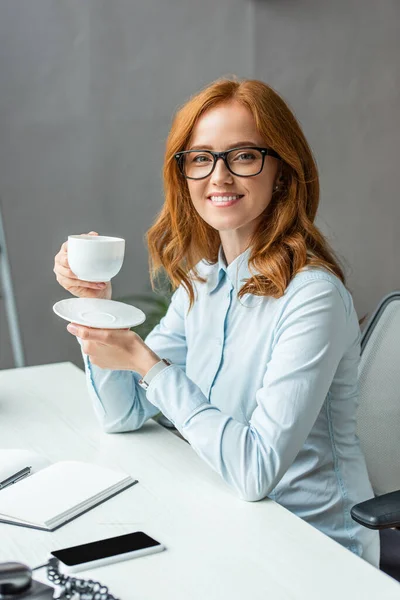 快乐的女商人 手里拿着咖啡杯和茶托 一边看着相机 一边坐在桌边模糊的背景上 — 图库照片