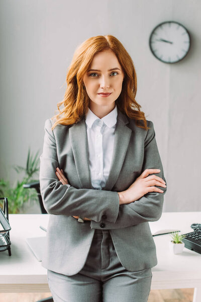 Front view of positive businesswoman with cross arms looking at camera, while leaning on table on blurred background
