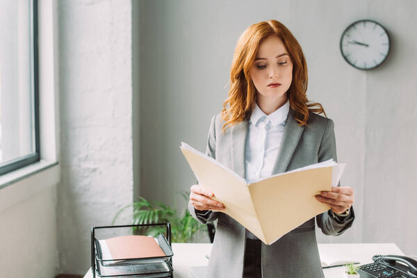 Serious businesswoman looking at folder with documents, while standing near workplace on blurred background