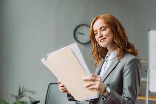 Smiling businesswoman looking at folder with paper sheets with blurred office on background