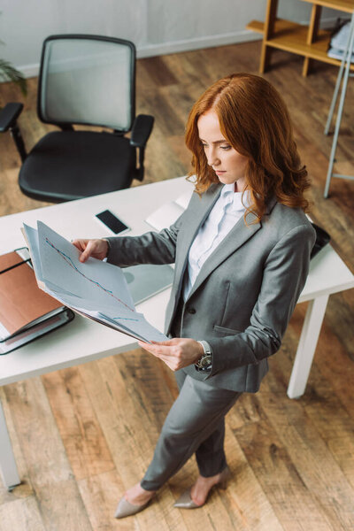 Overhead view of businesswoman looking at folder with graphs, while standing near workplace 