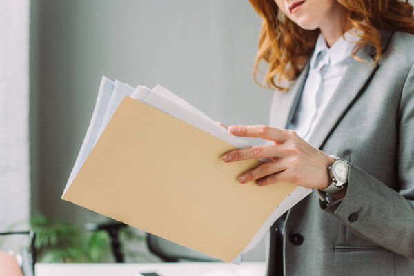 Cropped view of businesswoman leafing through paper sheets in folder with blurred workplace on background