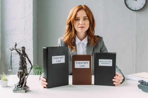 Redhead lawyer looking at camera and holding books with copyright law and intellectual property lettering, while sitting at table