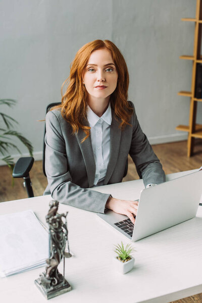 Redhead female lawyer looking at camera, while typing on laptop at workplace with blurred themis figurine on foreground