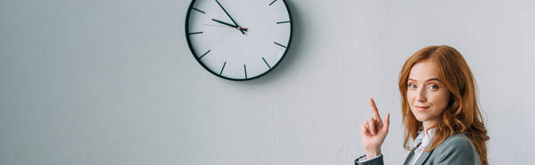 Позитивная рыжая деловая женщина, указывающая пальцем на настенные часы, глядя на камеру на сером, баннере