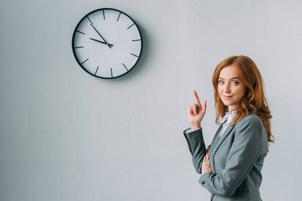 Smiling redhead businesswoman pointing with finger at wall clock, while looking at camera on grey