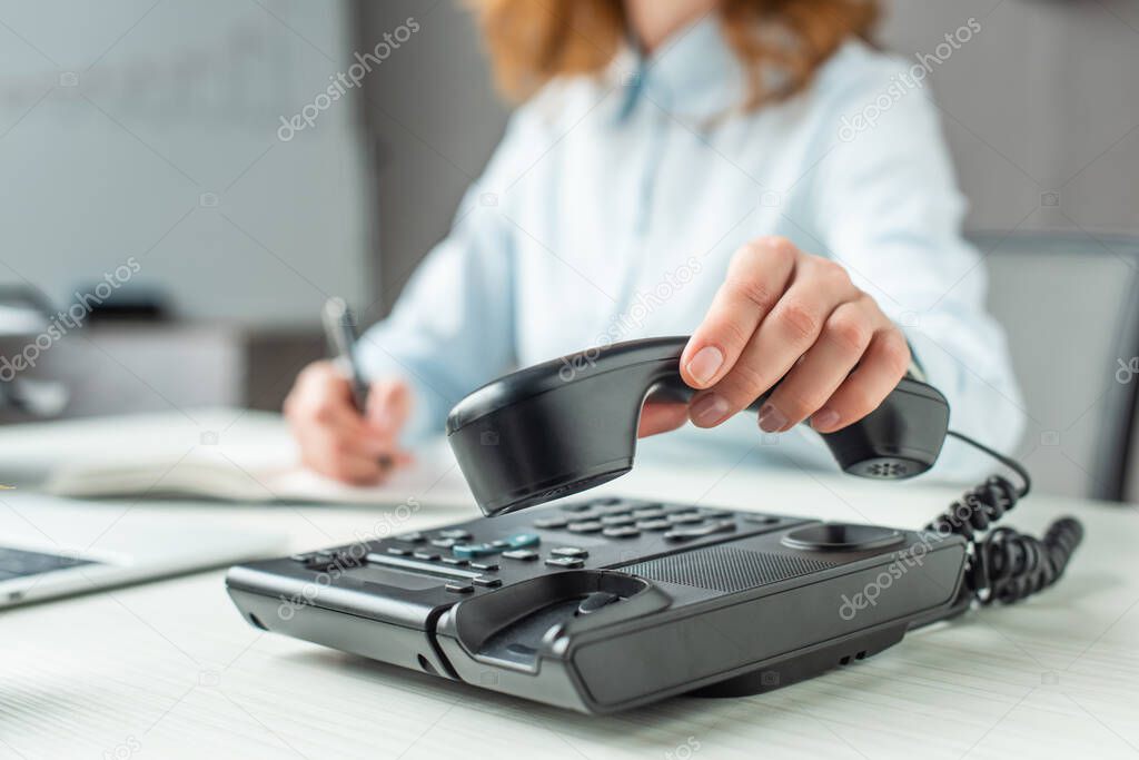 Cropped view of businesswoman putting handset on landline telephone at workplace on blurred background