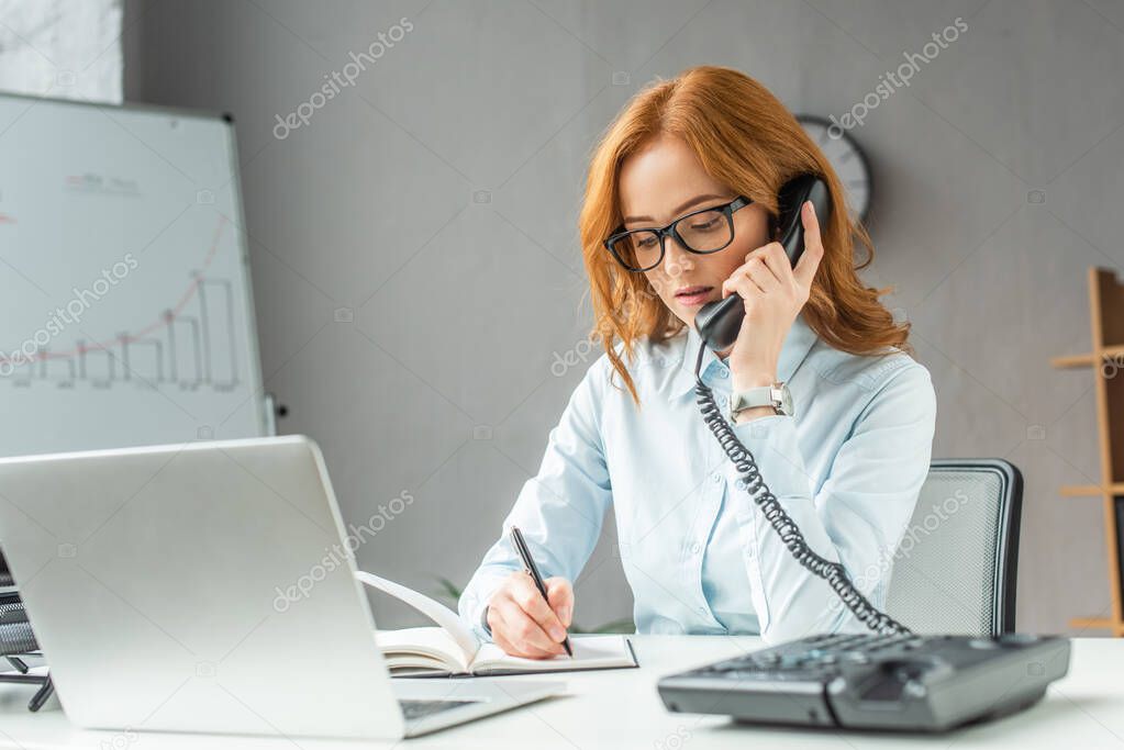 Redhead businesswoman talking on landline telephone, while writing in notebook at workplace on blurred foreground