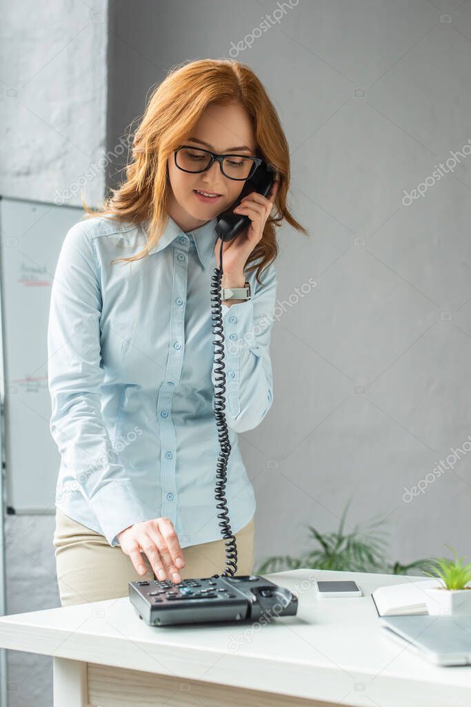 Positive businesswoman dialing number on landline telephone, while standing near workplace with digital devices 