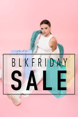 Yüksek açılı mavi kürk ceketli genç bir kadın alışveriş çantalarının yanında oturuyor ve pembe harflerle Cuma günü yanıp sönüyor.