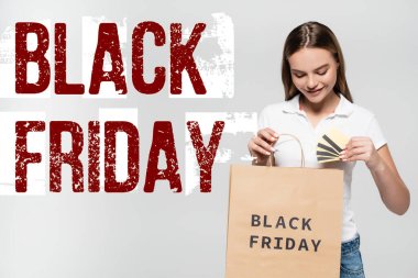 joyful woman holding credit cards and looking at shopping bag near black friday lettering on grey clipart