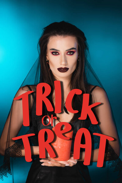 young pale woman with black makeup and veil holding burning candle near trick or treat lettering on blue