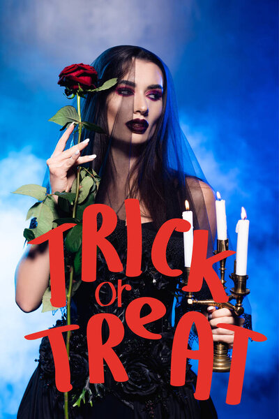 young woman in black dress and veil holding rose and burning candles near trick or treat lettering on blue with smoke